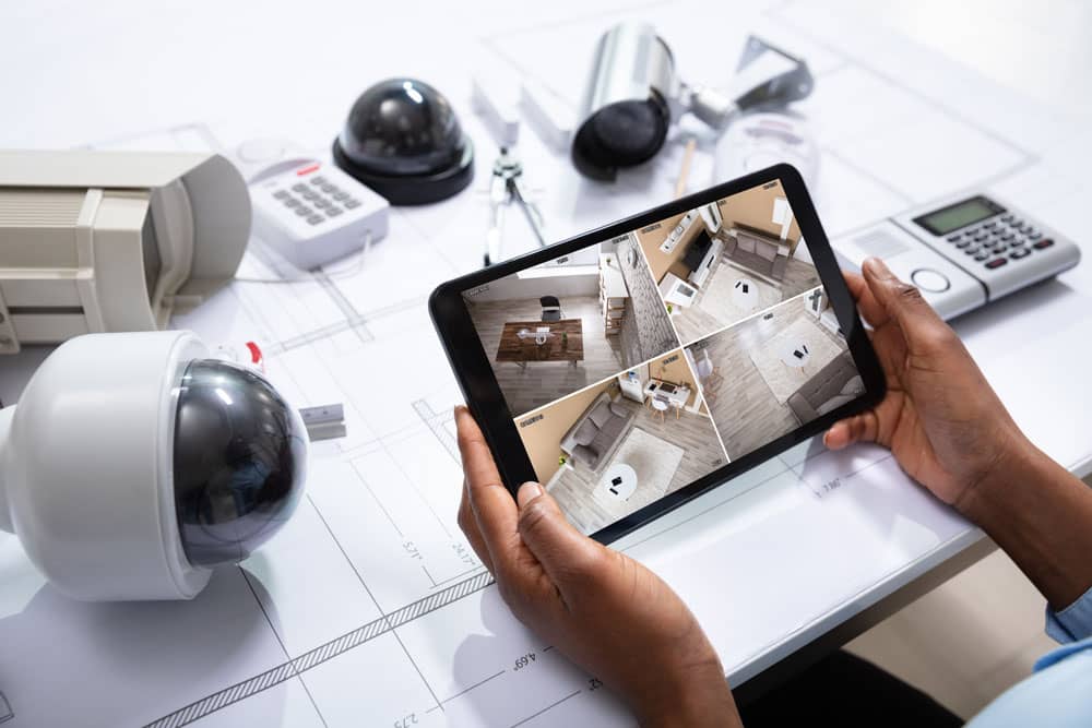 A person holds a tablet displaying a live feed from four security cameras. Surrounding the tablet on a desk are several security cameras, a calculator, and parts for security equipment. Blueprints are also visible underneath the equipment, possibly provided by CCTV dealers in Chennai.
