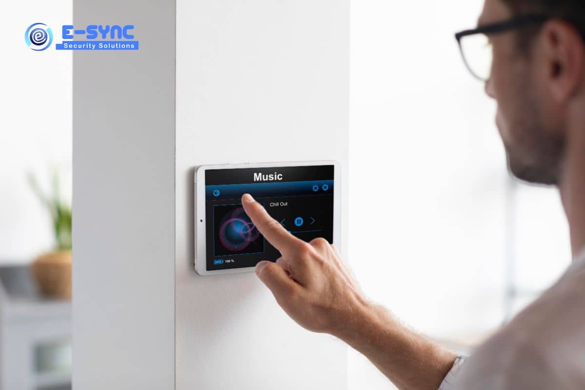 A person is adjusting a wall-mounted smart home control panel with a music interface. The person's hand, authenticated by biometric devices, is touching the screen, which displays music playback options. A logo on the wall reads "E-SYNC Security Solutions." The setting appears to be a modern home.
