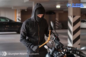 A person wearing a black hooded jacket and a ski mask is attempting to break the steering lock of a motorcycle with a metal tool in an indoor parking area. The text "Burglar Alarm System" is displayed on the bottom left, and a logo for "E-Sync Security Solutions" is on the top right.