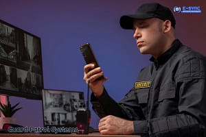 A security guard in a uniform and cap looks intently at his smartphone, utilizing Guard Patrol Systems. He sits at a desk with multiple monitors displaying surveillance footage. The background is softly lit in shades of blue and purple, and the logo "E-SYNC Security Solutions" is visible in the top right corner.