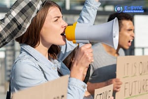 A woman shouts into a megaphone during a protest, using it as one of the many public announcement systems among the crowd. She holds a sign high, while other protesters with partially obscured signs stand behind her. The logo "E-SYNC Security Solutions" appears in the top right corner of the image.