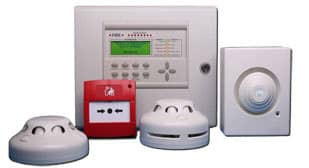 A security system setup is displayed with various components. Centered is a main control panel with buttons and a display screen. Surrounding it are two smoke detectors, a red fire alarm pull station, and another sensor device on the right.